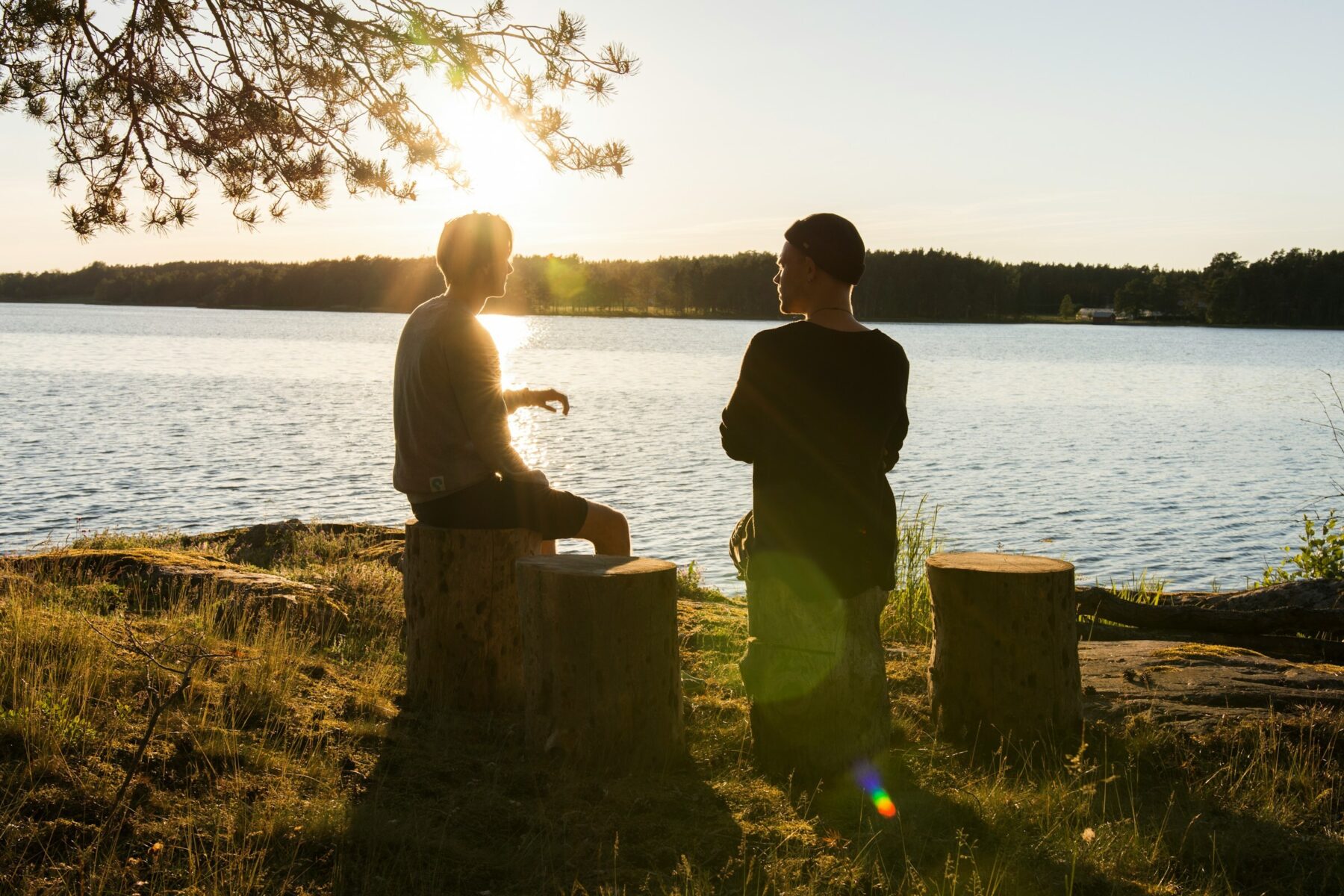 Two men sitting outside on tree stumps, beside a lake, in conversation