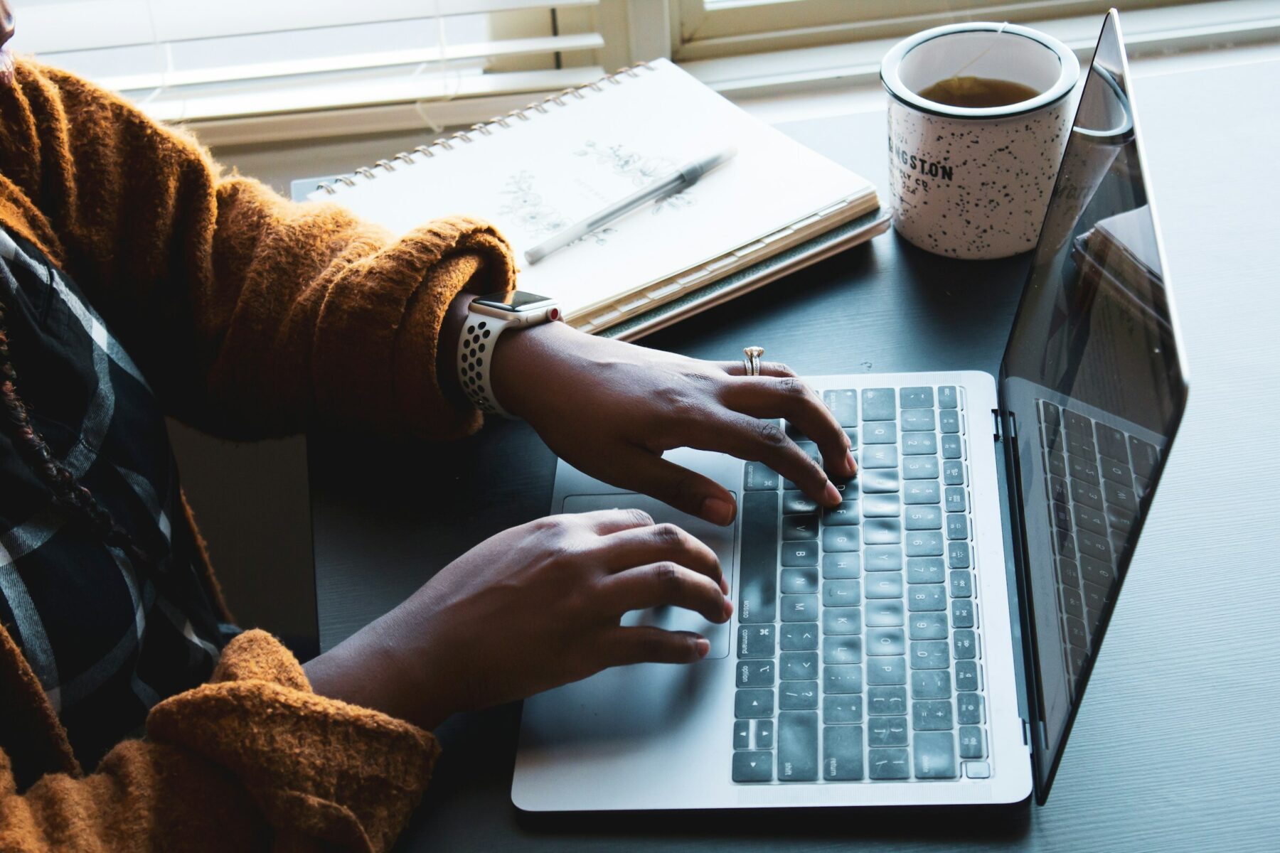 Close up image of a person typing on a laptop keyboard. There is a pen and paper, and a cup of tea, in the background