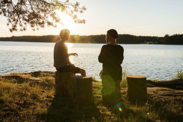 Two men sitting outside on tree stumps, beside a lake, in conversation